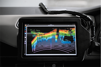 Fotografia de um monitor de tela no interior de um carro com a imagem de várias silhuetas de objetos em meio a vários feixes de cores azul, verde, amarelo e laranja.