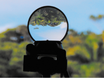 Fotografia de uma lupa com a lente evidenciando uma paisagem invertida, com o céu azul na parte inferior e vegetação na parte superior. Ao fundo, está a mesma paisagem vista na posição real e desfocada.