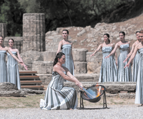 Fotografia de várias mulheres usando vestidos iguais, estilo tomara que caia, listrados e longos. Uma delas está no centro, agachada acendendo uma tocha de fogo em um espelho parabólico. Ao fundo há várias ruínas de pedras.