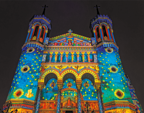 Fotografia da fachada de uma basílica vista de baixo para cima. Ela possui duas torres laterais e no centro, na parte inferior há três arcos, colunas no andar superior e uma parte triangular acima. Toda a fachada está repleta de desenhos coloridos, as torres têm cor predominante azul e verde com muitas estrelas amarelas. Na parte central há imagens de santos e predomina fundo azul, verde e estrelas amarelas.