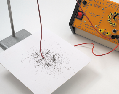 Fotografia de um fio magnético ligado a energia, passando de um lado ao outro de uma superfície retangular branca. Nessa superfície há partículas metálicas compondo um desenho circular em volta do fio.