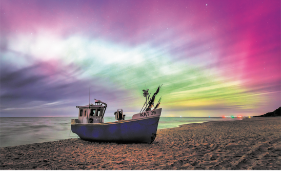 Fotografia de um barco parado na areia de uma praia. Há raios coloridos preenchendo o céu em tons de rosa, verde, amarelo, azul e roxo.