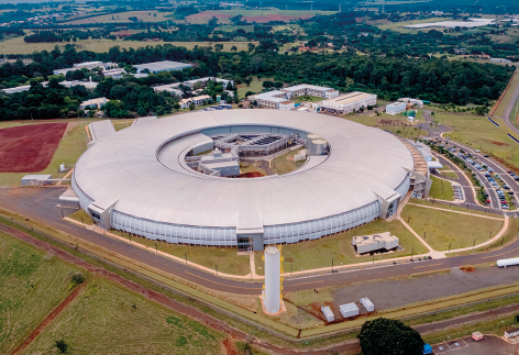 Fotografia da vista aérea de um acelerador de partículas. Ele é uma grande construção em formato circular, sendo semelhante a um estádio de futebol. Ao redor, há muitas áreas verdes, avenidas e outras construções.