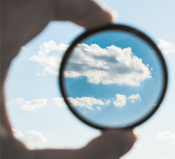 Fotografia de uma mão segurando uma lente em direção ao céu. A lente tem formato circular e através dela o céu é visto com mais nitidez, azul mais escuro e nuvens grandes e um pouco escuras. Fora da lente, o céu é visto em tons mais claros e com as nuvens também em cor mais clara.