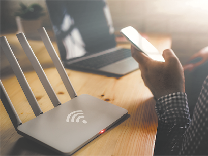 Fotografia de um modem de wifi sobre uma mesa e próximo de um notebook. Diante da mesa, há uma pessoa segurando um celular.