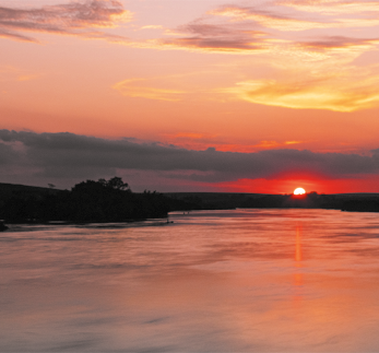 Fotografia de um rio com vegetação ao fundo e o sol se pondo no horizonte. Toda a paisagem é vista em tons de laranja que está mais forte ao redor do sol.