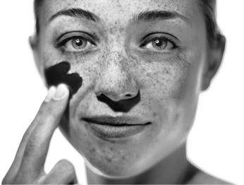 Fotografia em preto e branco com foco no rosto de uma mulher passando um produto líquido na bochecha. O produto tem cor preta e nas demais regiões do rosto dela há várias pequenas manchas escuras.