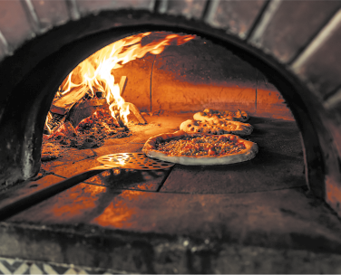 Fotografia de um forno à lenha contendo três pizzas. Ao lado das pizzas há bárias lenhas com chamas.