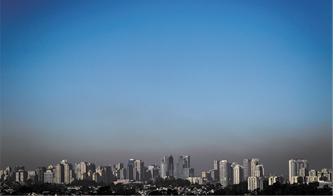 Fotografia vista de longe de uma cidade com vários prédios, construções e vegetação. Acima das construções o céu está azul, sem nuvens. Mais próximo dos prédios e construções há uma camada acinzentada na atmosfera.