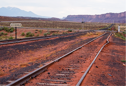 Fotografia de trilhos de trem em um caminho de terra. Uma das barras de ferro que compõe o trilho está deformada com uma leve parte curva em sua extensão. Ao fundo há montanhas e vegetação.
