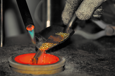 Fotografia de uma mão usando luva, segurando uma colher com pequenos pedaços de ouro. A colher está inclinada sobre um recipiente aquecido e com a cor alaranjada. Ao lado da colher há também parte de um cano com uma chama.