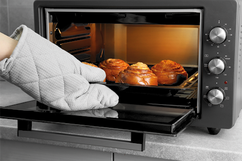 Fotografia de um forno elétrico com a tampa aberta. Há uma mão com luva térmica segurando uma bandeja com pães doces que estão dentro do forno.