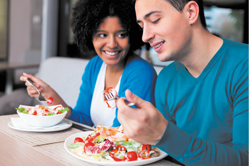 Fotografia de uma mulher e um homem sentados diante de uma mesa fazendo uma refeição. A mulher, vestindo uma blusa azul e camiseta branca, está olhando para o homem e segurando um garfo com um pouco de salada que está no prato, em frente a ela. O homem está olhando para o prato e segurando um garfo com um pouco da salada que está no prato em frente a ele.