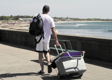 Fotografia de um homem vistos de costas, usando boné, camiseta e bermuda, segurando uma mochila nas costas e carregando uma grande caixa térmica com rodinhas. Ele está andado em uma calçada à beira mar. Ao fundo há uma praia, mar e vegetação.