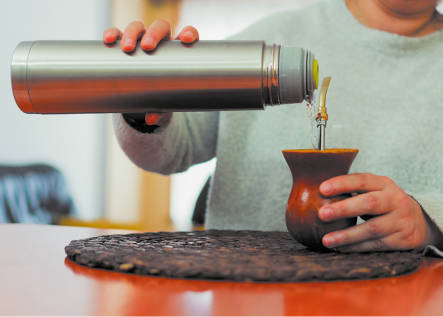 Fotografia de uma pessoa segurando uma garrafa térmica de metal que está inclinada e derramando água em uma cuia de chimarrão com um canudo de metal. A cuia está apoiada em uma mesa.