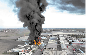 Fotografia com vista aérea de um incêndio em uma construção. A fumaça que sai do incêndio é escura e alta. Ao redor há várias outras construções industriais.