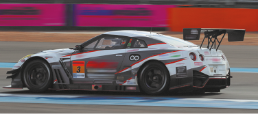 Fotografia de um carro de corrida em uma pista. Ele tem cor branca com cinza e vermelho, e na parte traseira há um aerofólio. Ao fundo, as proteções laterais da pista estão desfocadas.