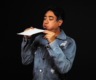 Fotografia de um homem segurando com as duas mãos uma tira de papel próximo da boca. Ela está assoprando a tira de papel, a qual está sendo levantada.