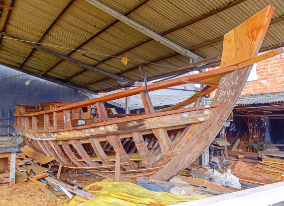 Fotografia em um barracão com uma construção de madeira da estrutura de um barco. Ao redor, há madeiras e materiais de construção.