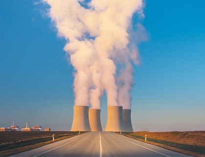 Fotografia de uma estrada e ao fundo, quatro chaminés de uma usina nuclear saindo fumaça branca. Nas laterais há vegetação, e mais ao fundo há algumas construções e o céu azul.
