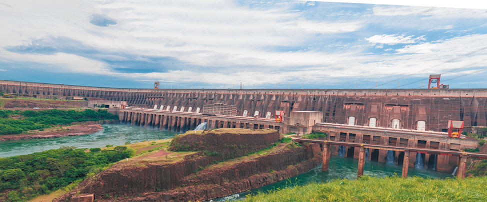 Fotografia de uma usina hidrelétrica. Há um rio passando por uma grande barragem. Nas laterais do rio há vegetação.