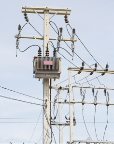 Fotografia de uma rede de energia elétrica, formada por um poste com fios elétricos na ponta, que são conectados a uma caixa.