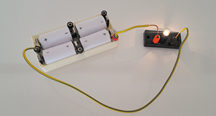 Fotografia de um circuito elétrico com uma caixa com 4 pilhas conectadas. Das laterais da caixa saem 2 fios que se conectam a uma estrutura pequena com uma lâmpada que está ligada.