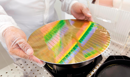 Fotografia das mãos de uma pessoa usando luvas e jaleco em um laboratório, segurando um disco com superfície metálica e quadriculada. No disco há o reflexo de faixas coloridas.
