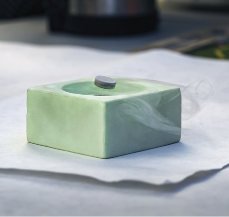 Fotografia de um bloco de cerâmica com a superfície concava. Nessa superfície há um pequeno imã com formato circular levitando sobre o bloco. Há fumaça em torno do bloco.