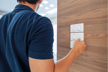 Fotografia de um homem visto de costas ao lado de uma parede. A mão do homem está apertando um botão do interruptor de luz que está na parede. Ao fundo há uma sala com várias luzes acesas.