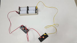 Fotografia de um circuito elétrico com uma caixa com 4 pilhas conectadas. Da lateral esquerda da caixa segue um fio vermelho que se conecta a um suporte com uma lâmpada. Do lado direito do suporte segue um outro fio vermelho que se conecta a um outro suporte com uma lâmpada. Do lado direito desse suporte segue um fio amarelo que se conecta ao lado direito da caixa com pilhas. As duas lâmpadas estão ligadas.