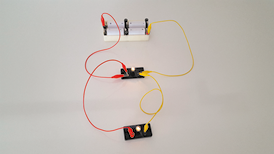Fotografia de um circuito elétrico com uma caixa com pilhas conectadas. Do lado esquerdo da caixa segue um fio vermelho que se conecta a um suporte com uma lâmpada. Do lado direito da caixa segue um fio amarelo que se conecta ao suporte com a lâmpada. Do lado esquerdo desse suporte segue um fio vermelho que se conecta a um outro suporte com uma lâmpada que está mais abaixo. Do lado direito há um fio amarelo que conecta os dois suportes com lâmpada. As duas lâmpadas estão ligadas.