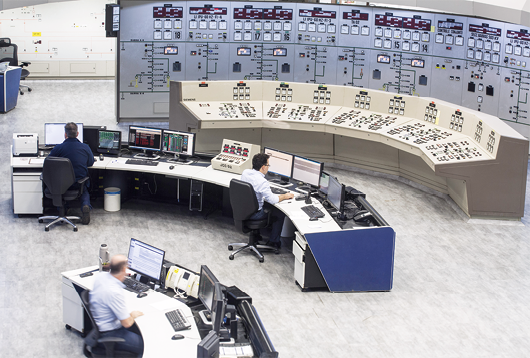 Fotografia de uma sala com uma grande máquina em uma parede. Ela tem vários controles e botões. Em frente a máquina há três mesas com controles, e pessoas usando computadores.