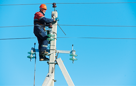 Fotografia de um homem no topo de um poste de energia elétrica. O homem está usando uniforme e equipamentos de segurança, incluindo capacete e luvas.