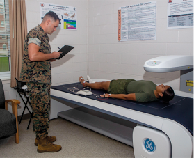 Fotografia de um homem em pé, usando uniforme do exército fazendo anotações com uma prancheta em um consultório. Em frente ao homem há uma máquina com uma maca, onde há uma mulher deitada de barriga para cima e braços alinhados ao corpo. Ela também usa uniformes do exército verde.