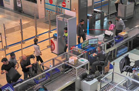 Fotografia com vista superior da área de embarque de um aeroporto. Há uma esteira com visão raio x para bagagens. Ao lado da esteira há pessoas passando por um detector de metal. E há pessoas retirando as malas da esteira.
