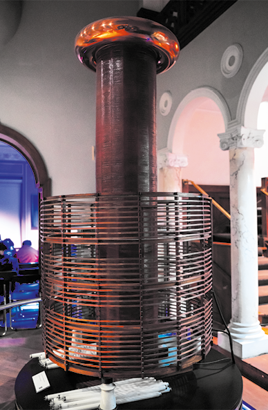 Fotografia de uma peça de metal com um grande cilindro no centro, com a parte superior arredondada e brilhante. Na base há uma grade em torno do cilindro. A peça está em uma sala de museu.