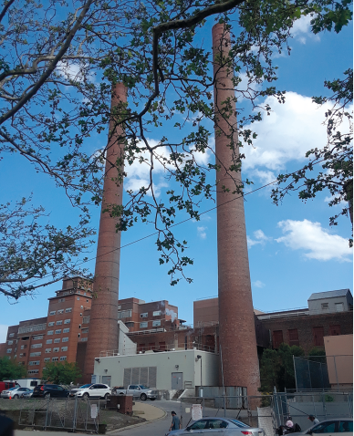 Fotografia de duas torres altas e estreitas, e entre elas há uma construção. Em torno das duas torres há outros prédios e um estacionamento com carros.