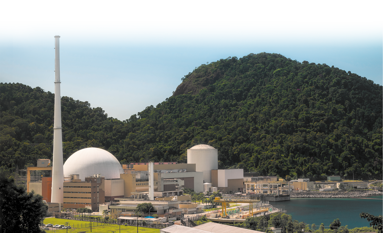Fotografia de uma usina nuclear. Há várias construções, uma delas tem formato esférico e algumas torres. Ao fundo há montanhas com vegetação.