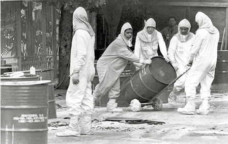 Fotografia em preto e branco de quatro homens vestidos com macacões brancos com capuz na cabeça. Eles estão segurando um barril de metal em um suporte com rodinhas. Mais à frente há um homem com macacão brando e capuz, olhando para os outros. E há mais barris de metal ao lado dos homens.