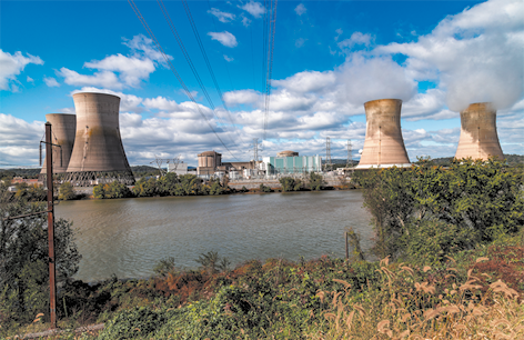 Fotografia de um rio com vegetação nas margens. Ao fundo há 4 chaminés largas de uma usina nuclear e construções entre as chaminés. O céu está azul e com nuvens.