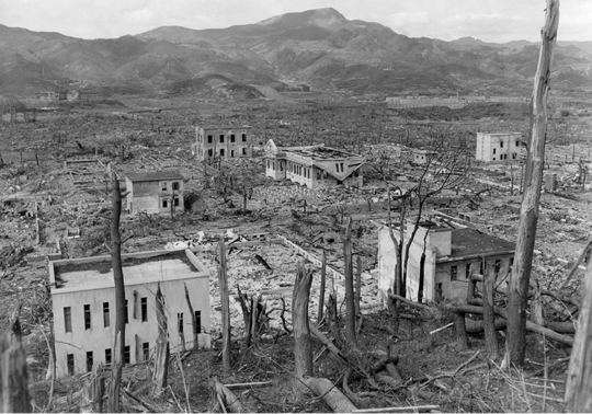 Fotografia em preto e branco de construções em meio a árvores queimadas e escombros. Ao fundo há montanhas.