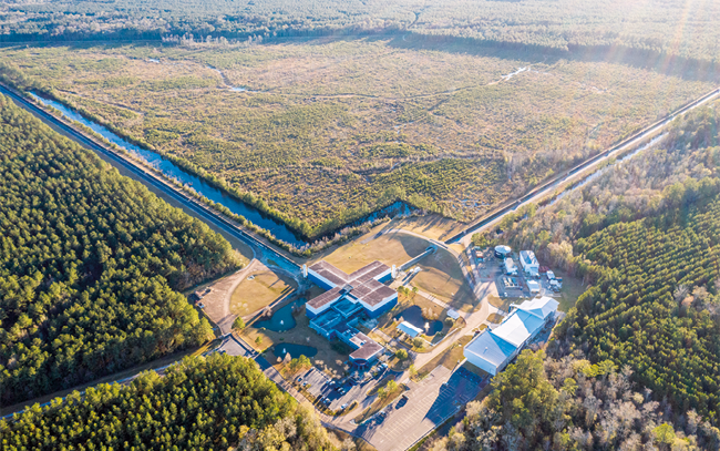 Fotografia com vista aérea de uma construção com vários galpões. Ela é cercada de plantações e há caminhos que seguem até essa edificação.