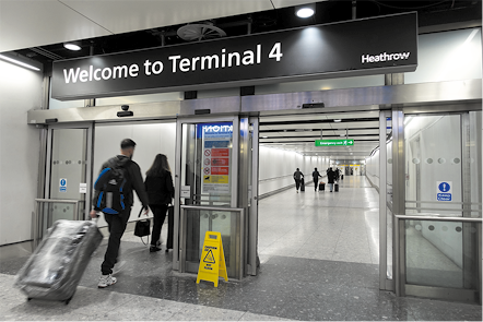 Fotografia de duas portas abertas, uma ao lado da outra, e pessoas passando por uma delas segurando malas. Acima das portas, há uma placa com a informação em inglês: welcome to terminal 4.