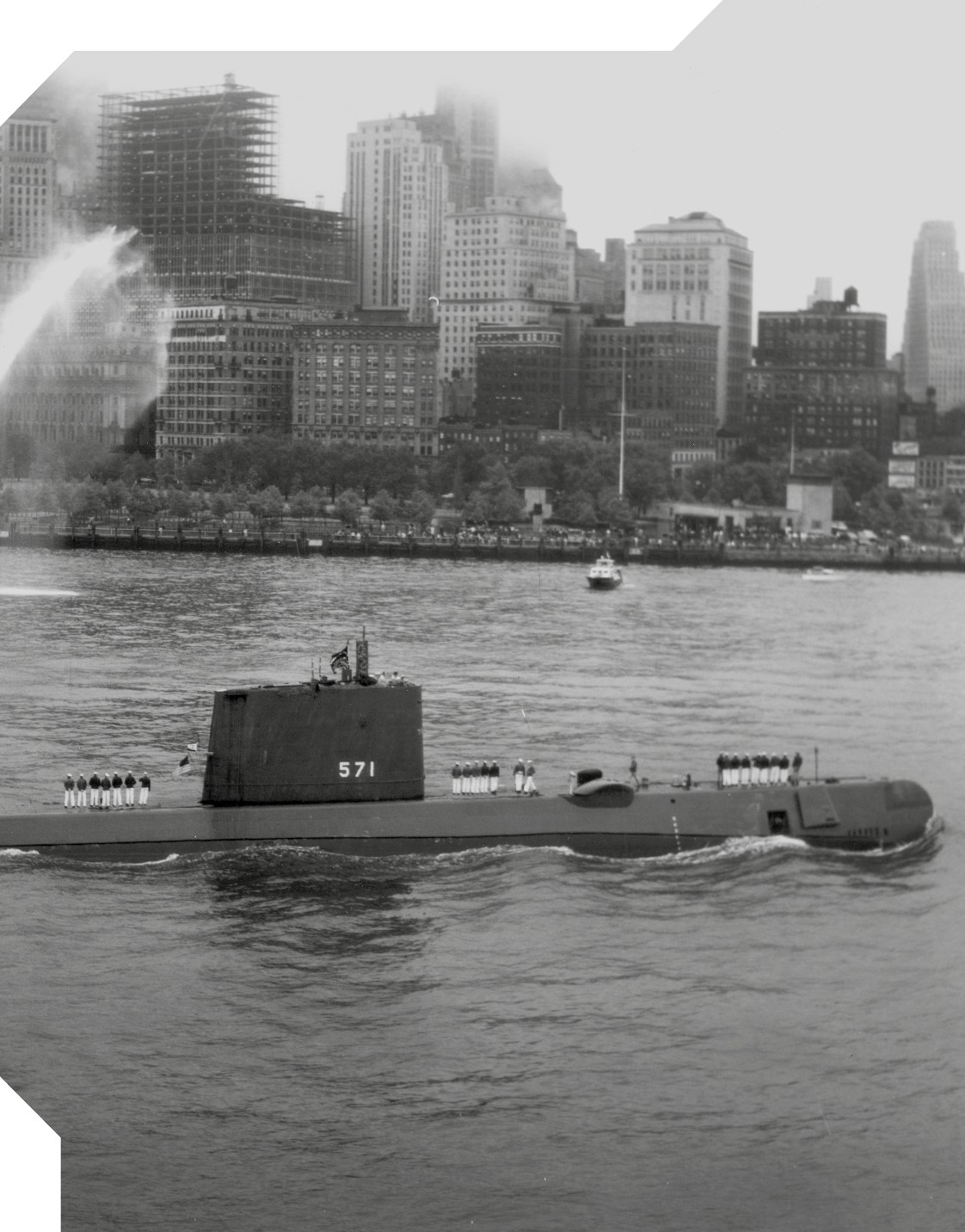 Fotografia em preto e branco de um submarino na superfície do mar, próximo as margens de uma cidade. Na cidade ao fundo há vários prédios e árvores.