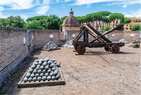 Fotografia de uma catapulta, com estrutura de madeira em formato retangular na base com quatro rodas, e acima há uma estrutura triangular e uma grande haste lateral. Ela está em um espaço cercado por um muro de tijolos e há várias pilhas de bolas de concreto ao redor.