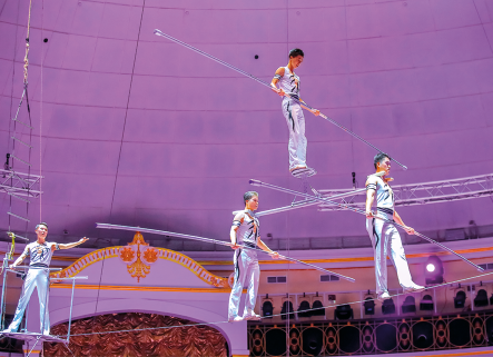 Fotografia de quatro homens usando roupas brancas e se equilibrando sobre cordas suspensas. Todos também carregam varas compridas.