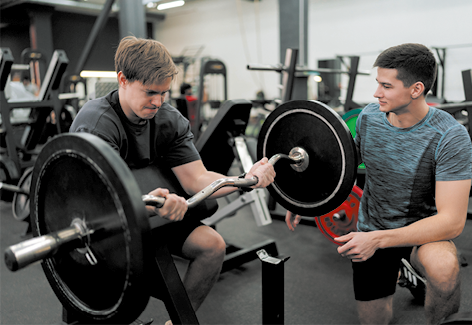 Fotografia de dois homens em uma academia. Um deles está sentado, levantando uma barra com pesos com os braços, mantendo os cotovelos apoiado. Ele apresenta expressão de força com os lábios bem fechados. O outro homem está ajoelhado ao lado, observando.