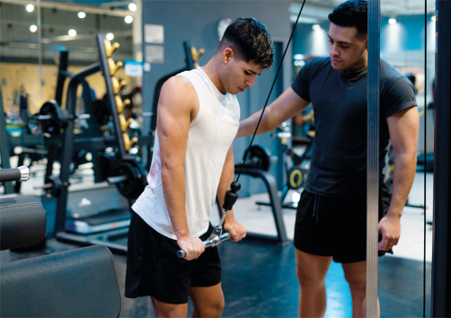 Fotografia de dois homens em uma academia. Um deles está em pé, diante de um aparelho composto por uma barra de ferro ligada a uma corda, a qual está empurrando na altura do joelho. O outro homem está logo ao lado, auxiliando com uma mão apoiada no braço do homem que faz o exercício.