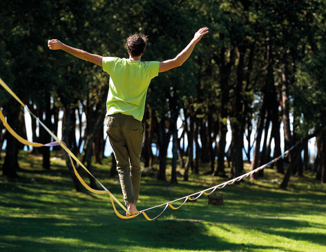Fotografia de uma pessoa vista de costas se equilibrando em uma fita suspensa do chão. Ela está em local gramado com muitas árvores.